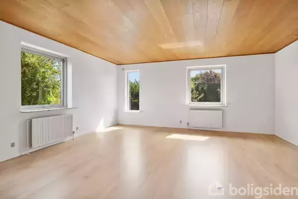 Et lyst rum med trægulv og træloft har tre vinduer med udsigt til grønne omgivelser. Rummet er tomt og har to radiatorer under vinduerne.
