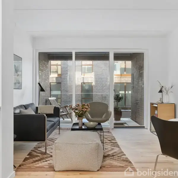 En stue med moderne møblering, herunder grå sofa, lænestole, og sofabord. Store vinduer giver udsigt til en altan med planter. Rummet er lyst og stilfuldt indrettet.