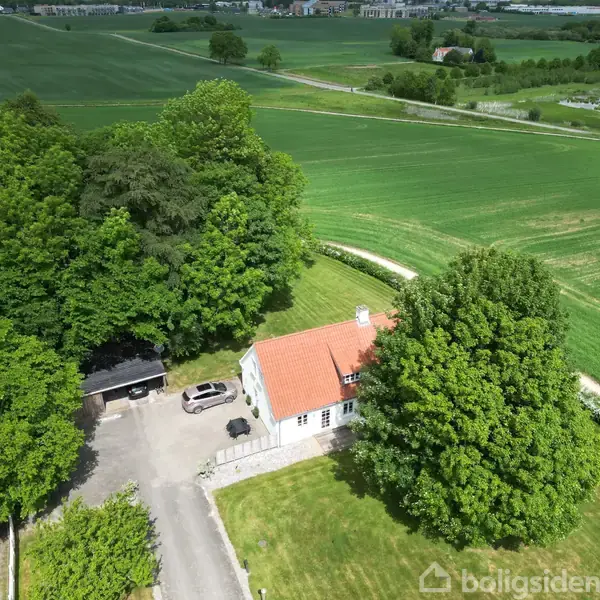 Hus med rødt tag står omgivet af grønne træer og marker. To biler er parkeret ved indkørslen, og en grusvej løber gennem landskabet.