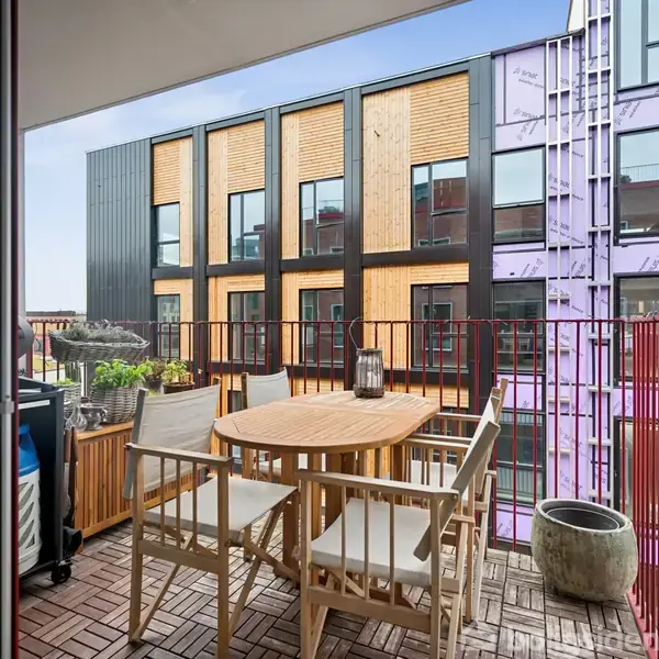 Rundt havebord med stole står på en altan med træfliser. Udendørs plantekasser og en grill ses omkring bordet. Baggrunden viser en moderne bygning med gul og lilla facade.