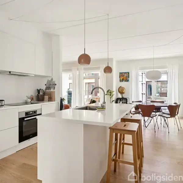 Køkken med hvidt interiør og ø, hvorpå der står to træstole; i baggrunden ses spiseplads med træstole og lyskuglelamper. En plante og udsmykning på væggene.