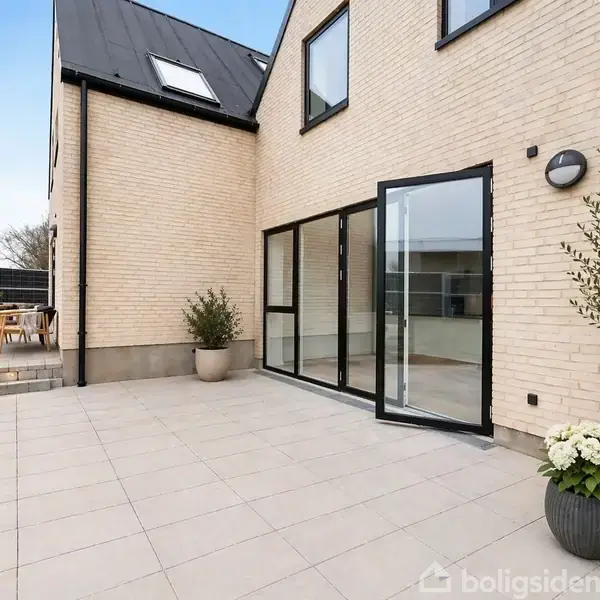Et moderne hus med lyse mursten har en rummelig flisebelagt terrasse, dekoreret med planter i krukker. Foran en stor glasdør står et bord med grill i baggrunden.