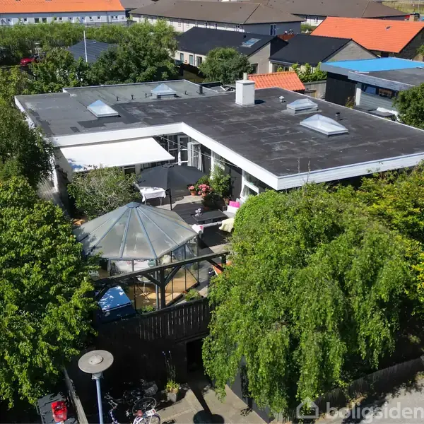 Et fladt taghus med tagvinduer omgivet af træer. En glaspavillon er synlig i haven. Nabohuse med farverige tage omkranser området. Der er parkerede biler ved huset.
