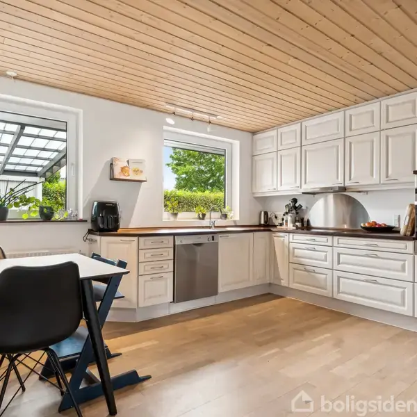 Et lyst køkken med træloft, hvide skabe og moderne apparater. Et spisebord med stole står ved siden af vinduer, hvor planter er placeret. Udsigt til en grøn have.