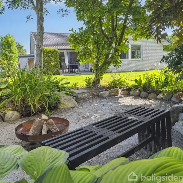Bænk står i have, omgivet af frodig vegetation og småsten. I baggrunden ses et hus med terrasse.