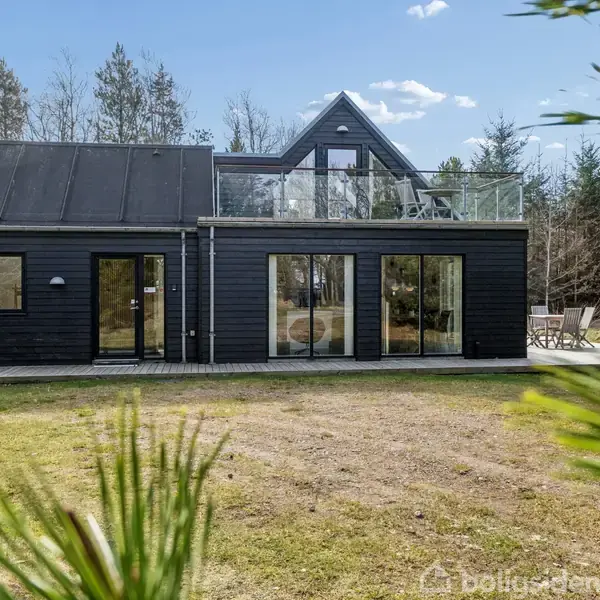 Sort træhus med store vinduer står stille i en grøn have. Terrassen ovenpå har glasrækværk. Omkring huset ses høje træer under en lys blå himmel.