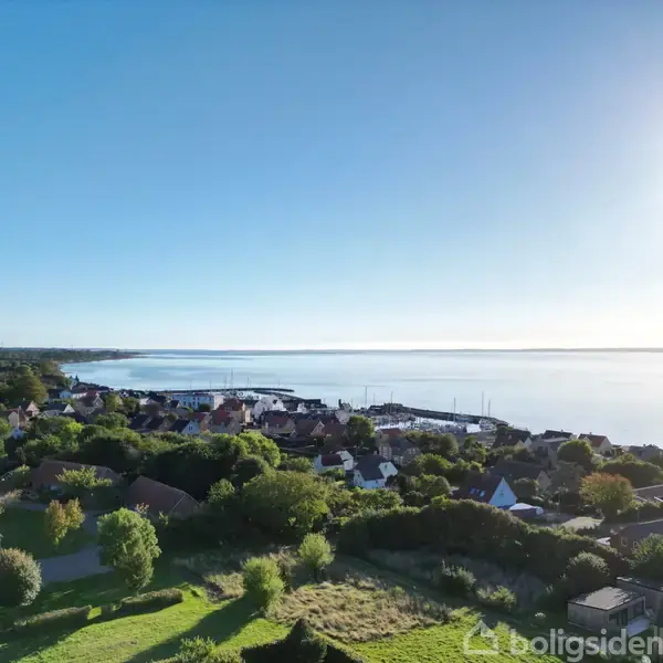 Bebyggelse med røde tage ligger blandt grønne træer og marker i et skovrigt landskab. Enkeltstående huse ses til venstre, omkranset af hække og velplejede haver.