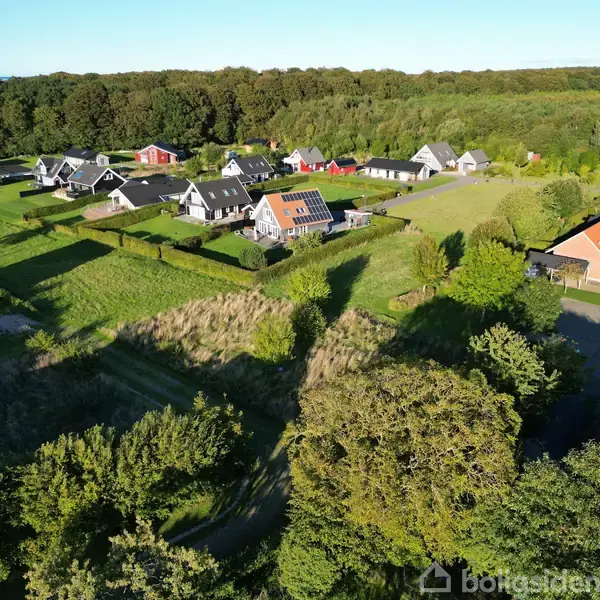Kystby med huse, træer og marina. Sollys skinner på stillestående vand. Huse spreder sig langs kystlinjen. Grønne områder omkring husene. Ingen tekst i billedet.