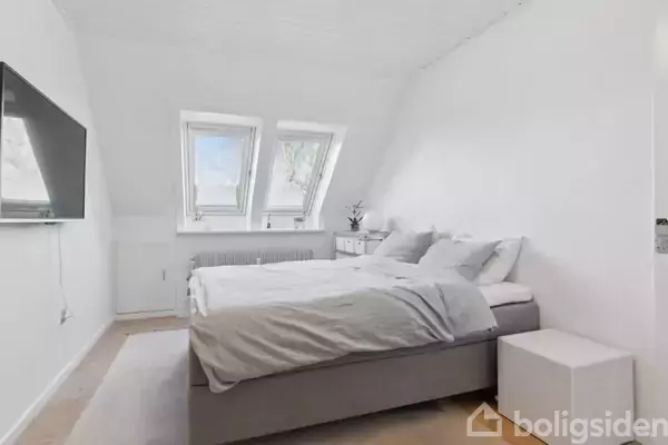Seng med gråt sengetøj står i et lyst soveværelse. To ovenlysvinduer giver naturligt lys. Fladskærms-tv hænger på væggen overfor sengen. Rummets indretning er minimalistisk.