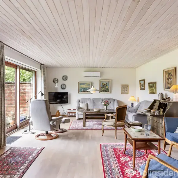 Stue med møbler og tæpper, indeholdende sofaer, lænestole og et sofabord. Væggene er dekoreret med malerier, og der er store vinduer mod en murstensvæg udenfor, hvilket skaber en lys atmosfære.