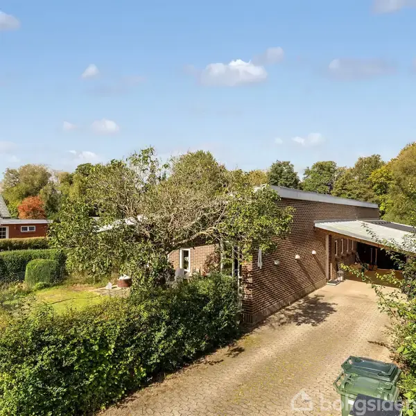 Murstenshus set bagfra i en grøn have, omgivet af buske og træer. Huset har en carport og en flisebelagt indkørsel under en klar himmel med lette skyer.