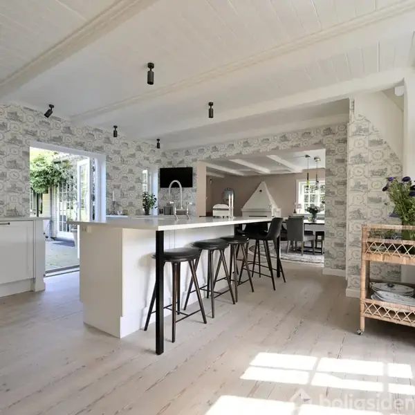 Køkken-alrum med køkkenø og barstole; lyse trægulve og tapet med mønster; i baggrunden en spiseplads ved store vinduer, der slipper lys ind, ved siden af en trappe.
