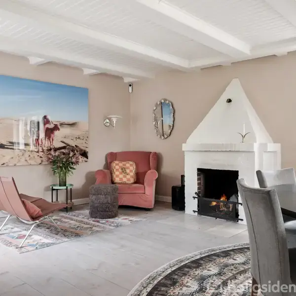 Stue med åben pejs, sofa og lænestol omkring et rundt tæppe. Væggen har et maleri af en strandscene. Et rundt spisebord står i højre side ved et vindue.