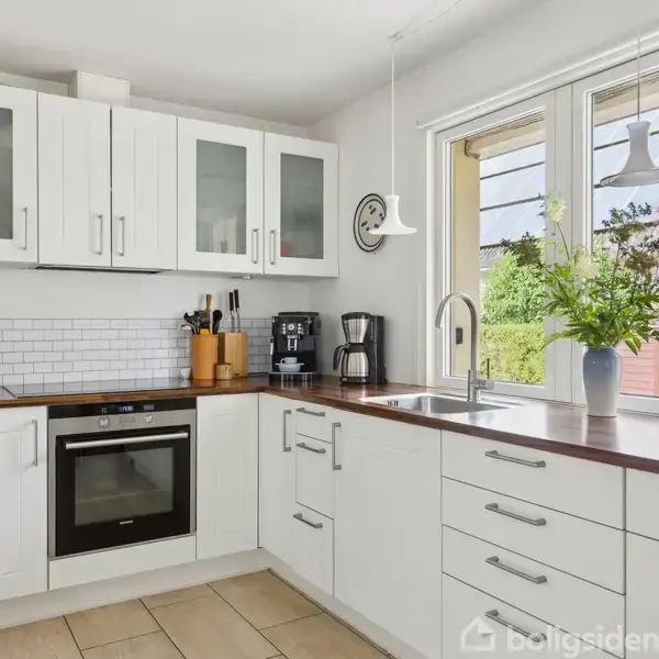 Hvidt køkken med indbyggede skabe og skuffer, kaffemaskine og ovn. Et vindue med udsigt over en have, en vase med blomster, og lysindfald gennem vinduet.