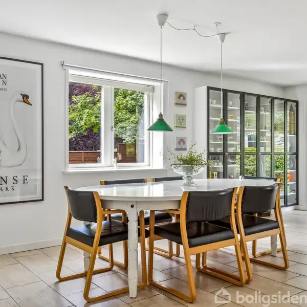 Et hvidt spisebord med seks læderstole står i en lys spisestue. En plakat med teksten "Hans Christian Andersen, 2 April 1805 - 4 August 1875, Odense, Denmark" hænger ved siden af et vindue.