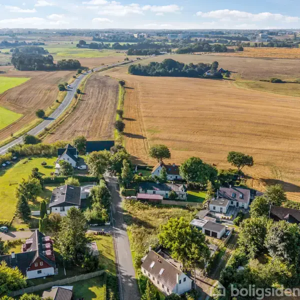 Et luftfoto viser en lille landsby omgivet af landbrugsmarker og veje. Husene ligger tæt sammen, omringet af træer, og i baggrunden ses flade landskaber og spredt vegetation.