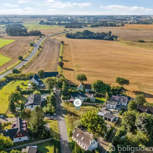 En åben grund med vildtvoksende græs og buske omgivet af træer. I baggrunden ses et hvidt hus blandt tæt bevokset skov. Himlen er klar med få skyer.
