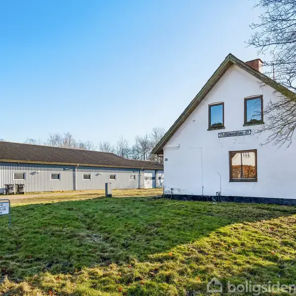 En hvid bygning med skilt "Tidligere Lægehus" står på en grøn græsplæne ved siden af en større lagerbygning og en vej. Et skilt i forgrunden har teksten "Til Salg Bymægler".