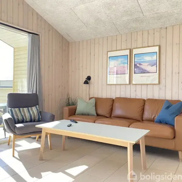Brun lædersofa med puder står foran en væg med to billeder. En lænestol med pude står ved siden af. Et sofabord er foran sofaen. Store vinduespartier giver lys.