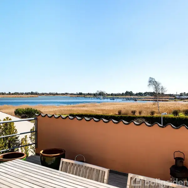 Terrasse med træbord og stole, stille står i forgrunden. Udsigten viser en sø eller fjord omkranset af græs og træer under en blå himmel. Ingen tekst er fundet.