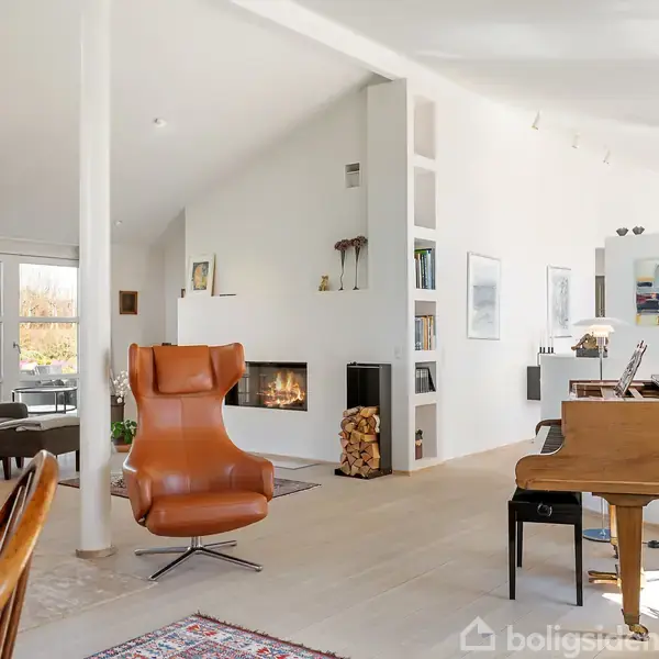 Brun lænestol står foran en pejs i en elegant, moderne stue med hvide vægge, høj loftshøjde og store vinduer. Et flygel er placeret ved siden af en spiseplads med hængende lamper.