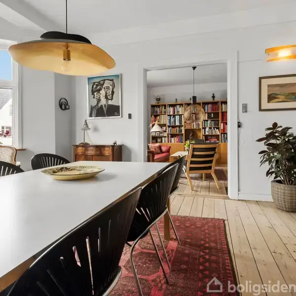 Spisebord med sorte stole står på et tæppe i et lyst rum. Væggene har kunst og planter, mens en døråbning viser en stue fyldt med bøger og en sofa.
