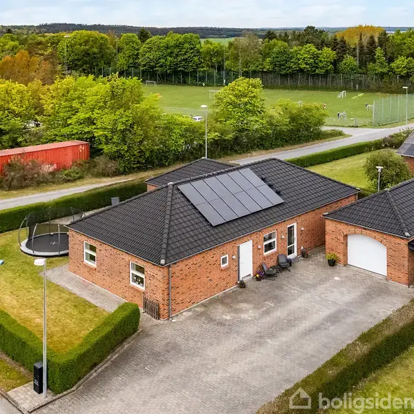 Et rødmurst hus med solpaneler på taget står i en velplejet have med trampolin og legetårn. En indkørsel med fliser ligger foran, omgivet af grønne områder og vej.