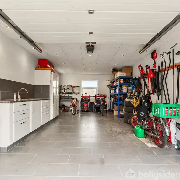 Garage med værktøjer og cykler opbevaret langs væggene. Vask og skabe til venstre, flere redskaber hængt til højre, med værktøjsreoler bagerst og flisebelagt gulv.