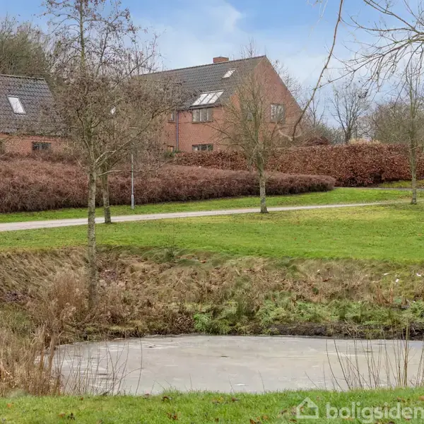 En lille dam ligger i forgrunden omgivet af græs og træer. I baggrunden ses to røde murstenshuse med sorte tage under en klar blå himmel.