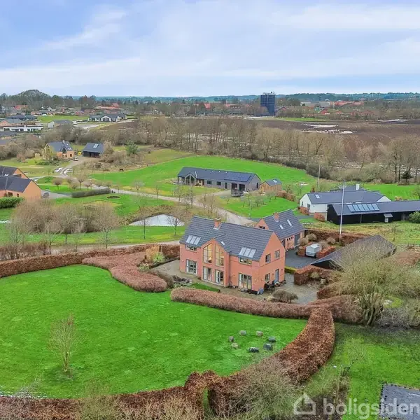 Hus med sort tag omgivet af grønt græs og hække. Beliggende i landligt område med flere spredte huse, marker og træer i baggrunden.