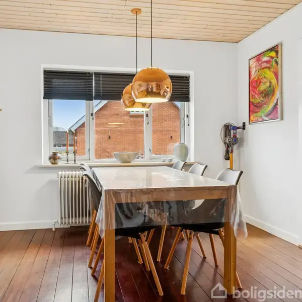 Et spisebord dækket med en gennemsigtig dug står midt i et lyst rum med træloft. Tre stole er placeret omkring bordet, og en kobberlampe hænger over det.