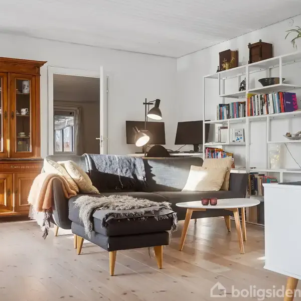 En sort sofa med puder og tæpper står i stuen foran en hvid bogreol fyldt med bøger. Ved siden af er et brunt vitrineskab, og på gulvet et rundt sofabord.