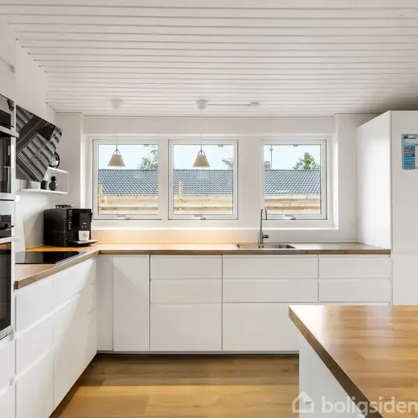 Et moderne køkken med hvide skabe og træbordplader. Køleskabet står til højre, mens ovn og kaffemaskine findes på venstre væg. Tre vinduer giver udsigt til tage.