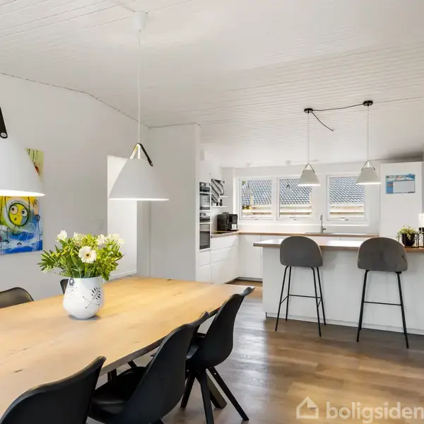 Langt spisebord med sorte stole, dekoreret med en blomsterbuket, står i en lys spisestue. Bagved ses en åben køkkenbar med høje stole og moderne indretning.