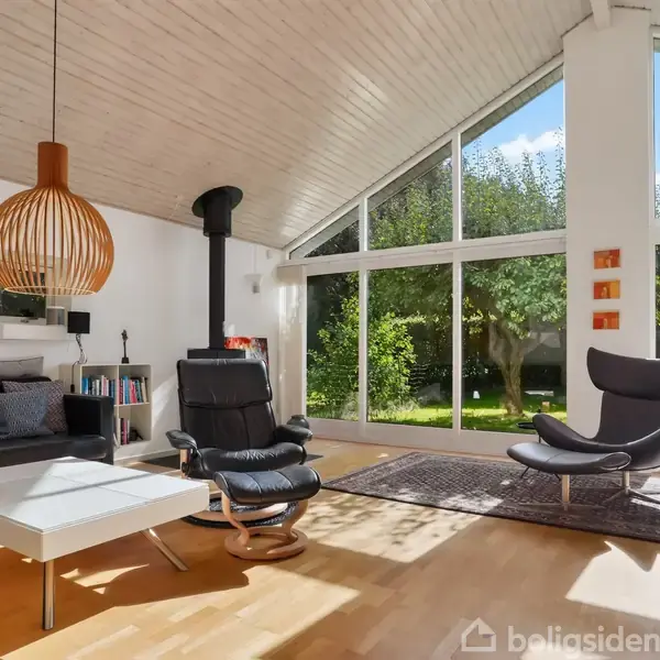 En moderne stue med lænestole og sofa på et lyst trægulv. Store vinduer giver udsigt til en grøn have udenfor. Skulpturel lampe hænger over bordet.