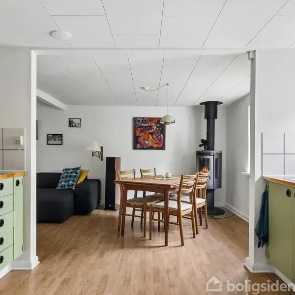 Køkken-alrum med grønne skabe og træbord omgivet af stole. Pejs i hjørnet og sort sofa i baggrunden. Vægge er dekoreret med billeder, og der er trægulv.
