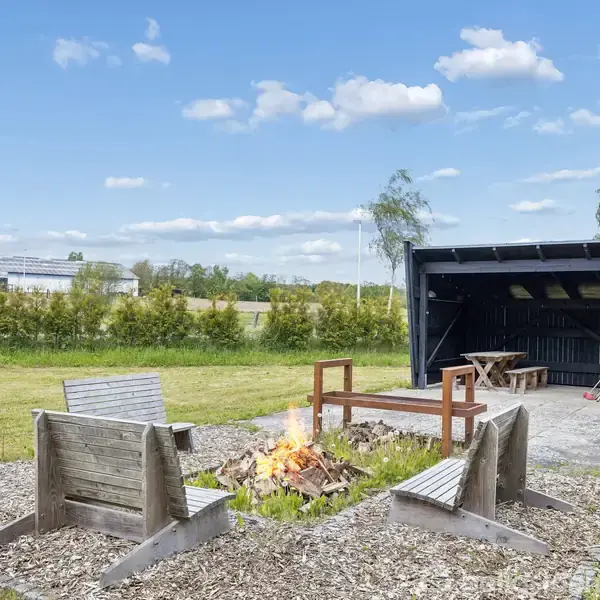 En rød murstensbygning med et skråtag står i grønne omgivelser. Ved siden af huset er en stor stabel brænde og nogle affaldsbeholdere. Området er frodigt og landligt.