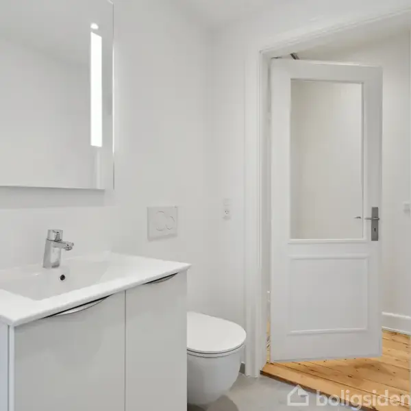 Hvid badeværelsesenhed med vask og spejl over, fastgjort i et moderne, minimalistisk badeværelse. Hængende toilet ved siden af. Åben dør fører til rum med trægulv.