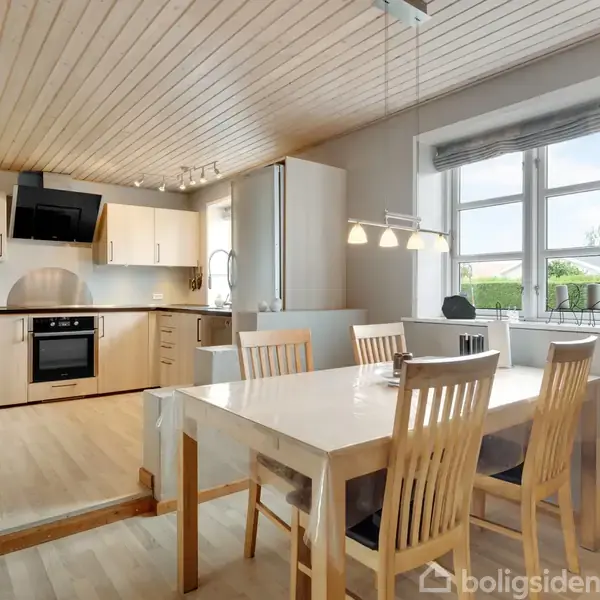Et moderne køkken med lyse træelementer har et spisebord med fire stole. Loftet er træbeklædt, og et stort vindue giver udsigt til grønne omgivelser.