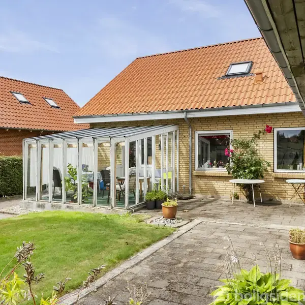 En gul murstensvilla med rødt tegltag omfatter en glasudestue med udsigt til en velplejet have. Terrasse med potteplanter og et bord, omgivet af hække og tilstødende bygninger.