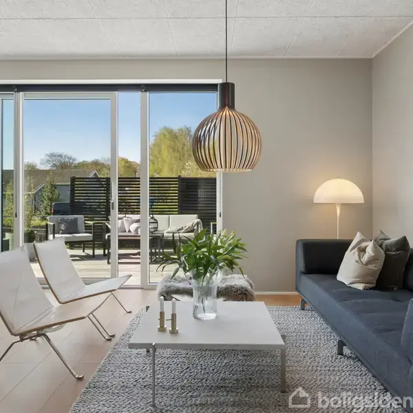 En moderne stue med en sort sofa, to hvide stole og et hvidt sofabord. En stor lampe hænger over bordet. Glidende døre åbner til en terrasse med udsigt over naturen.