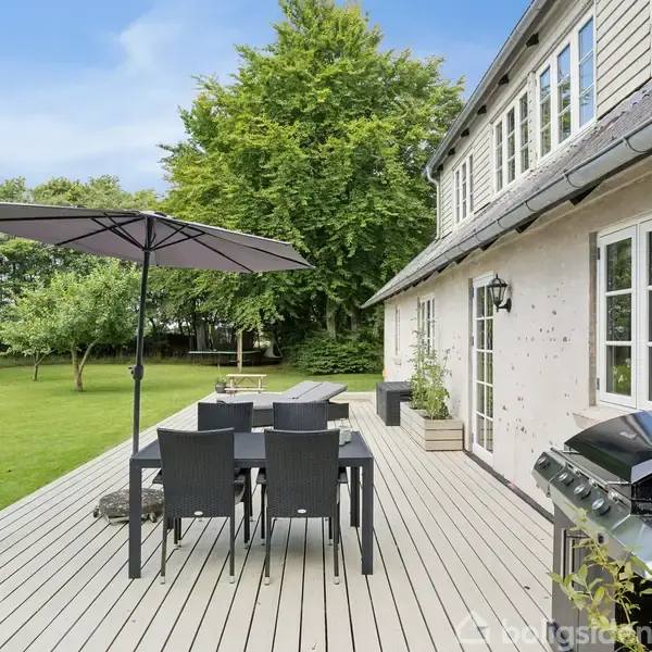 En terrasse med havemøbler og parasol står foran et hus med store vinduer. En grill placeret ved siden af terrassen. Baggrunden viser en grøn have med træer.