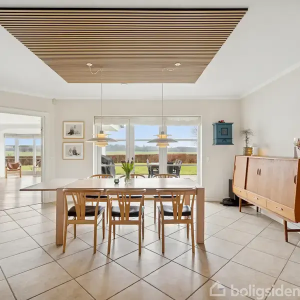 Et spisebord med træstole står centralt i et lyst rum med store vinduer og flisegulv. Et skab ved siden af, og en åben dør fører til en anden stue.