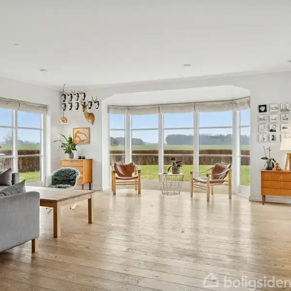 Stue med gråt sofaarrangement og lænestole ved vinduer. Bordlampe og plante skaber hygge. Store vinduer giver udsigt til natur. Trægulve og minimalistisk indretning.