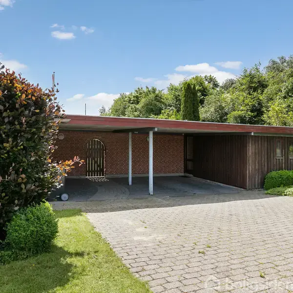 Carport i træ står ved siden af et rødt murstenshus, omkranset af en brostensbelagt indkørsel og grønne buske; himlen er klar med få skyer.
