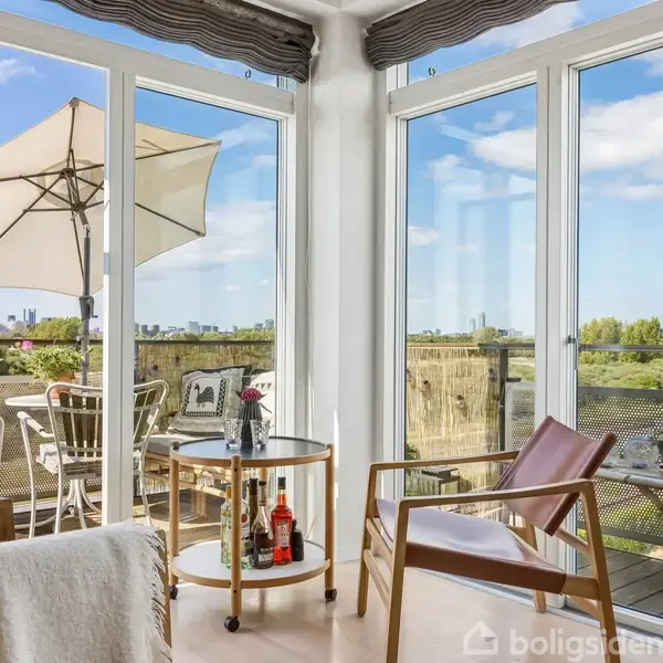 To lænestole står omkring et lille bord med flasker og glas. Et vinduesparti viser en altan med parasol og stole, med udsigt til grønne træer og byens skyline i baggrunden.