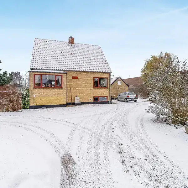 Et gult hus dækket af sne står stille i en villavej med buske og parkerede biler omkring. Sneen dækker også vejen og haven.