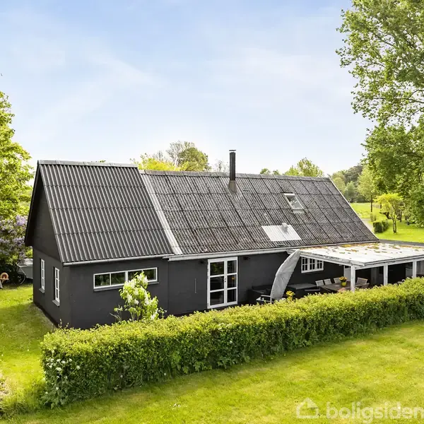 Et sortmalet hus står i en grøn have omkranset af buske. Taget har en mørk farve, og en del af huset er overdækket med et terrasseområde.