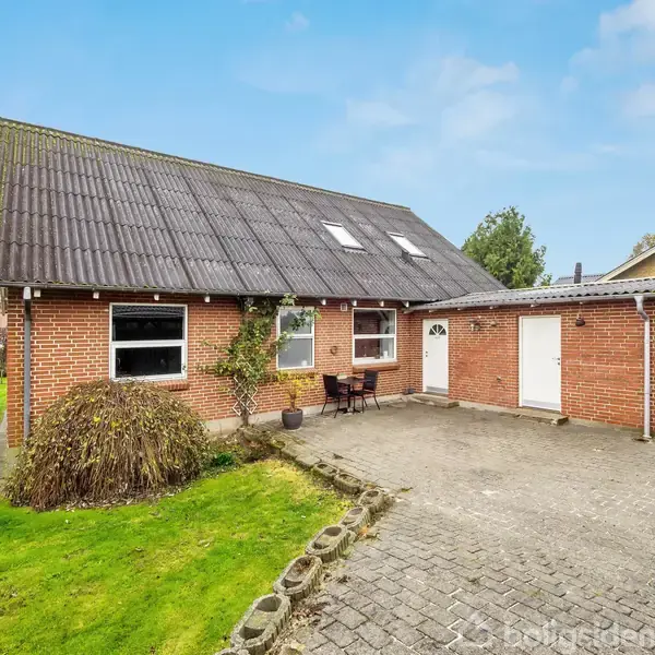 Et rødt murstenshus står stille på en velplejet græsplæne, omgivet af andre huse. Foran huset er en flisebelagt indkørsel og et lille bord med stole til afslapning.