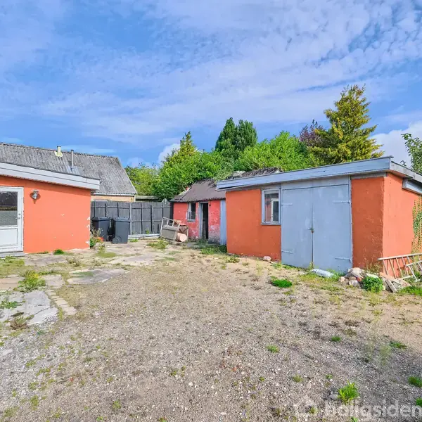 Rødt udhus står stille i en solbelyst gårdsplads omgivet af forskellige planter og buske. Flere bygninger med lysegrå tage ses i baggrunden under en blå himmel.